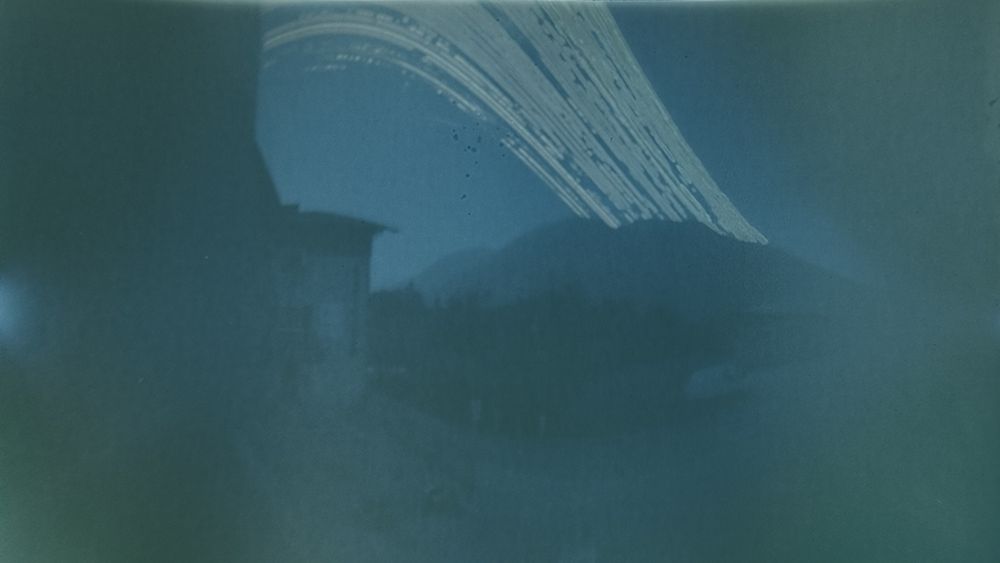a two-tone image showing multiple arcs of the sun's path at the top. The edge of a building is just about visible in the foreground, and mountains with a clear ridge curtail the sun's path at various points