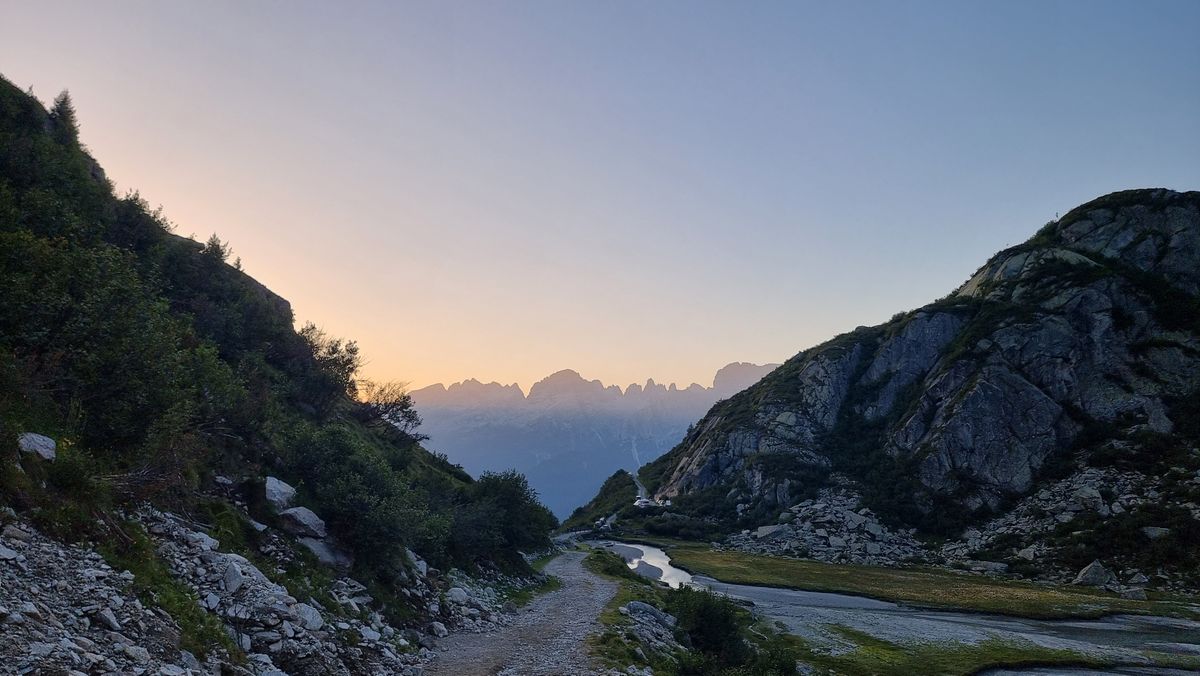 dawn view of a jagged mountain ridge on the horizon framed by rocky and partially vegetated valley sides in the nearfield. In the foreground a gravel track descends from the camera. Lower down to the right is a river and with a small grassy bank. The sky is pale blue and there aren't any clouds.