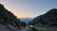 dawn view of a jagged mountain ridge on the horizon framed by rocky and partially vegetated valley sides in the nearfield. In the foreground a gravel track descends from the camera. Lower down to the right is a river and with a small grassy bank. The sky is pale blue and there aren't any clouds.