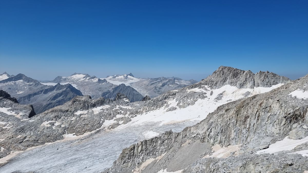 A high-altitude panorama with rocky peaks and small glaciers as far as the eye can see, all under a bright blue sky
