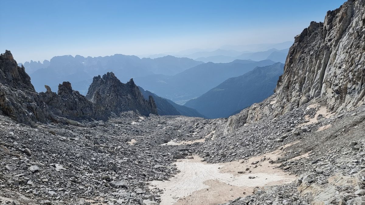 a debris covered glacier is framed by rocky walls on either side. In the distance are a series of ridges and valleys silhoutted. the sky is clear and blue.