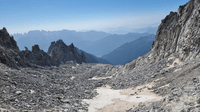 a debris covered glacier is framed by rocky walls on either side. In the distance are a series of ridges and valleys silhoutted. the sky is clear and blue.