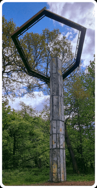 A sculpture in the woods: a cement and steel tower, with an irregular hexagon of rusty steel atop, like a halo, set in woodland
