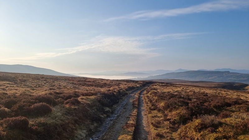 a track leads across a brown heather covered hillside. A few ridgelines visible in the distance. The sky is blue. In the distance, within a small valley the top of a cloud inversion is just visible